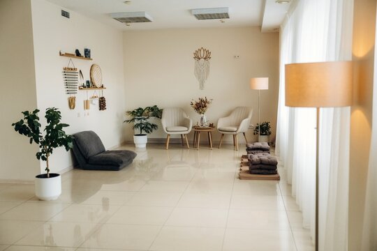 Bright modern living room with cream chairs, gray lounge seat, indoor plants, folded blankets, pendant wall decor, and glossy tile flooring in a contemporary home interior