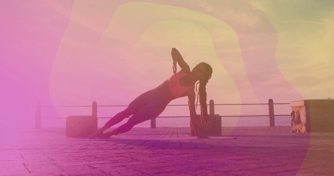 Holding side plank, woman in pink sports bra and leggings balancing on promenade, with mat, railing
