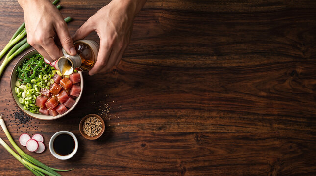 Overhead flat lay of chef bare hands drizzling sesame oil on tuna poke bowl