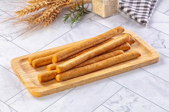 Wooden board holding baked breadsticks with rosemary and wheat stalks on marble table surface