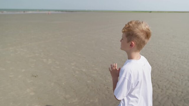 A boy is walking along a wide sandy beach slow motion playing with sand in his hands