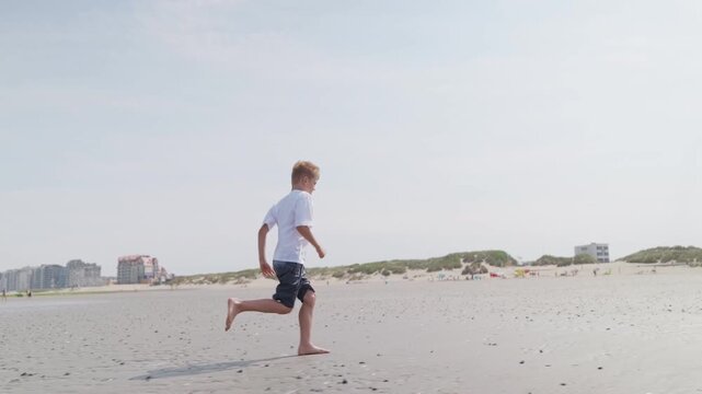 Boy running along wet beach sand dunes and buildings in background slow motion by the sea