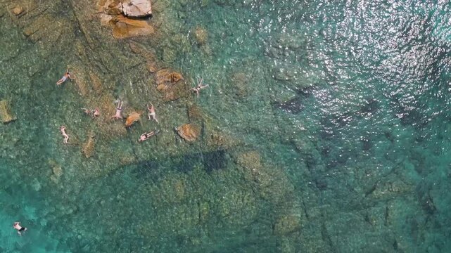 An overhead view of crystal clear, turquoise waters along a rocky coastline, with people snorkeling.