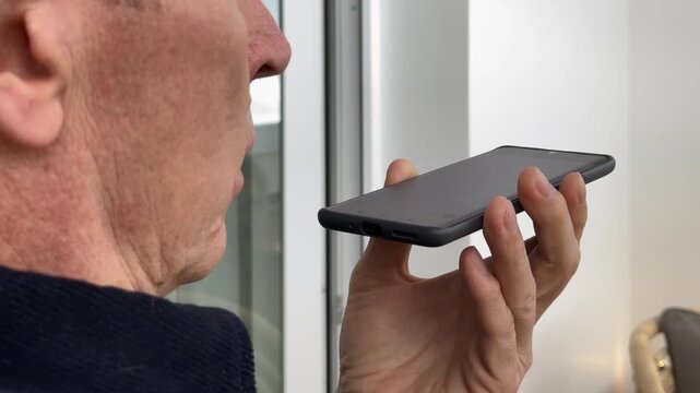 Close up of man holding phone at his mouth, voice chat with AI assistant. Speaking a question to smartphone, mature old man. Audio message at home.