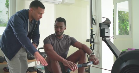 Male client and trainer coaching while grey-shirted client rowing on rower at home gym near weights © WavebreakMediaMicro