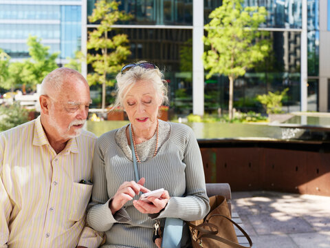 Senior woman using smart phone and talking to elderly man while sitting in city park