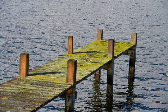 Verwitterter Holzsteg mit gelbgr&uuml;nem Moos f&uuml;hrt &uuml;ber dunkelblaues, leicht bewegtes Wasser, w&auml;hrend Sonnenlicht die raue Holzstruktur und Wasserreflexionen hervorhebt.