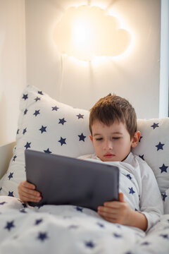 Cute Little Boy Lying in Bed Watching Digital Tablet Under Glowing Night Lamp