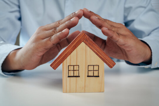 Businessman hands making roof shape gesture over wooden house model on white desk, concept of home insurance, property protection, family safety, and real estate investment.