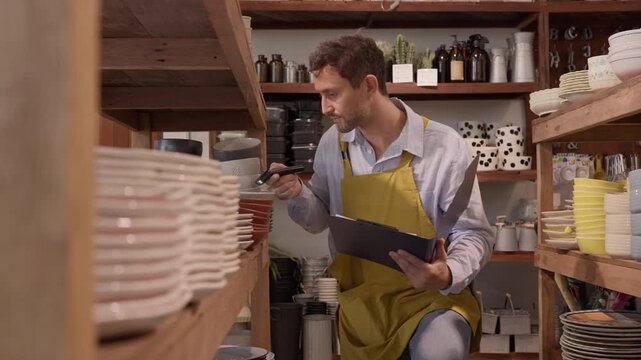Ceramic Shop Worker Using a Handheld Scanner and Clipboard to Check Plate Inventory, Caucasian Male Employee Reviewing Merchandise on Retail Shelves