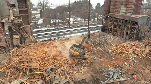 An excavator moves broken wood and debris between factory structures beside railway tracks on an overcast day, with patches of snow visible beyond the site.