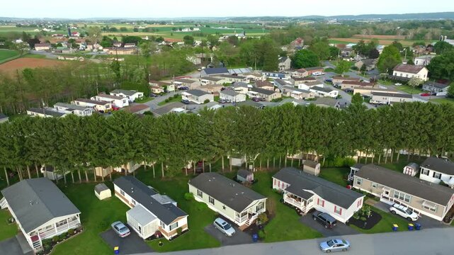 Aerial shots show a neighborhood with several mobile. manufactured houses, parked cars, and open fields. The surroundings include trees and rural land. The area appears organized with clear roads.