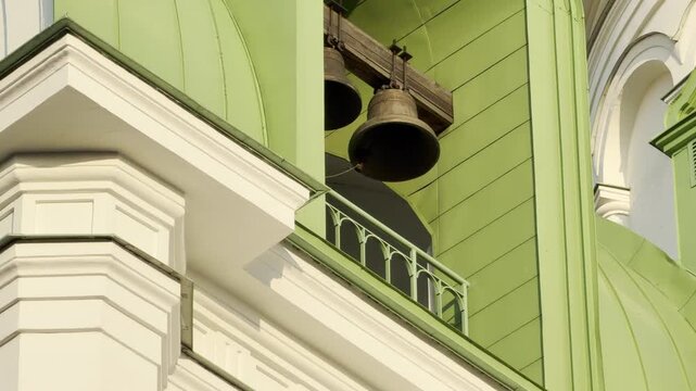 A heavy bronze bell hangs in the green architectural tower of an Orthodox cathedral in Pushkin, Russia. The rhythmic swinging creates deep, resonant chimes that echo solemnly through the air.