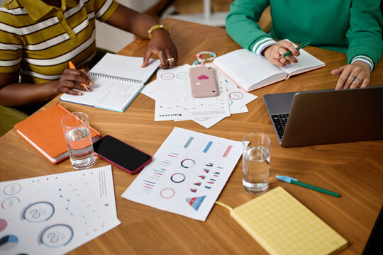 Diverse young adult women analyzing data charts and taking notes at coworking desk with laptop. Suitable for corporate teamwork and business analytics marketing