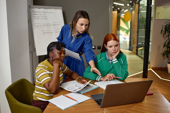 Multiethnic group of young adult businesswomen collaborating over laptop and documents in modern coworking office. Visual represents corporate teamwork, suitable for marketing campaigns