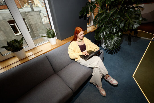 High angle view of young adult Caucasian woman working on laptop in modern coworking office. Suitable for freelance career, remote employment, digital workflow marketing campaigns