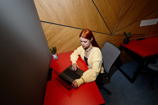 High angle view of young adult Caucasian woman working on laptop in modern coworking office, ideal for remote work, freelance technology, and modern corporate productivity campaigns