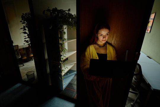 Young Caucasian woman working overtime on laptop standing in dark coworking office doorway. Useful for cybersecurity, deadline management and remote tech advertising