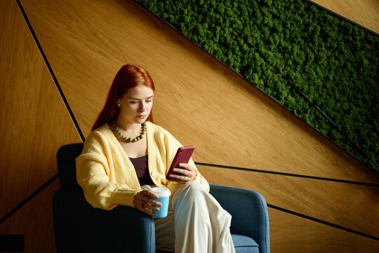 Young Caucasian woman using smartphone holding coffee cup in coworking office. Scene suitable for corporate mobile communication, digital lifestyle marketing, remote work advertising