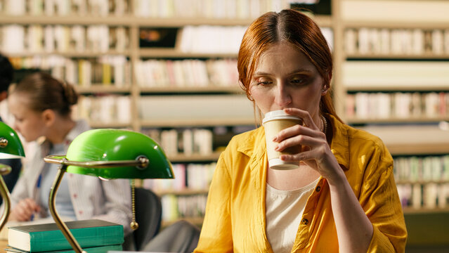 Student, coffee and library as woman drinks takeaway beverage at desk beside books and green lamp. Concentration, balance and independent learning support academic routines and study breaks.