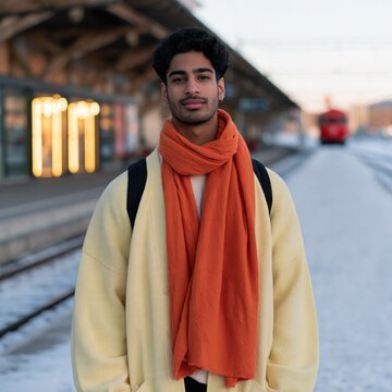 Young man standing on a snowy train platform wearing an orange scarf and pale coat for winter travel poster and social media background