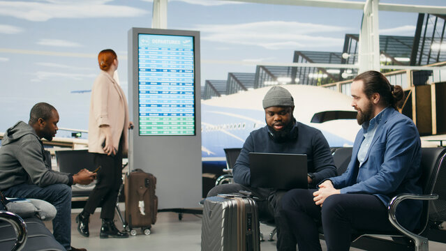 Airport colleagues, laptop and discussion as two male travelers talk beside suitcases in departure lounge. Business mobility, teamwork and coordination support professional tasks between flights.
