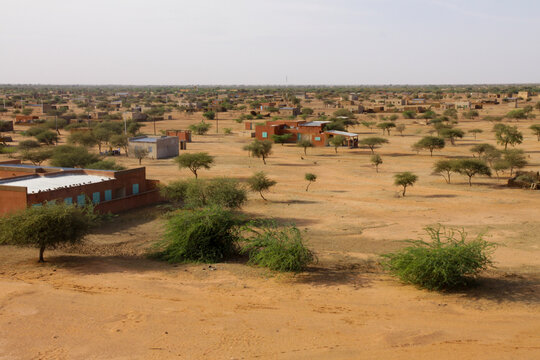 Person walking across a sandy plain with acacia trees and buildings, Sahel Region, Markoye, Burkina Faso
