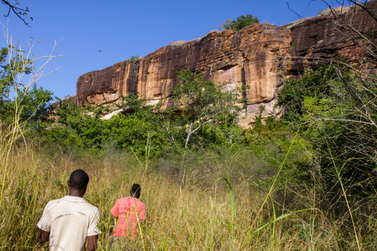 Two people walking in tall grass toward the cliff of the Abandoned Troglodyte Village, Cascades Region, Niansogoni, Burkina Faso
