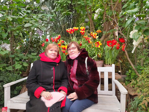 Two older women sit together on a white bench among flowering plants, enjoying companionship during a visit to a lush greenhouse