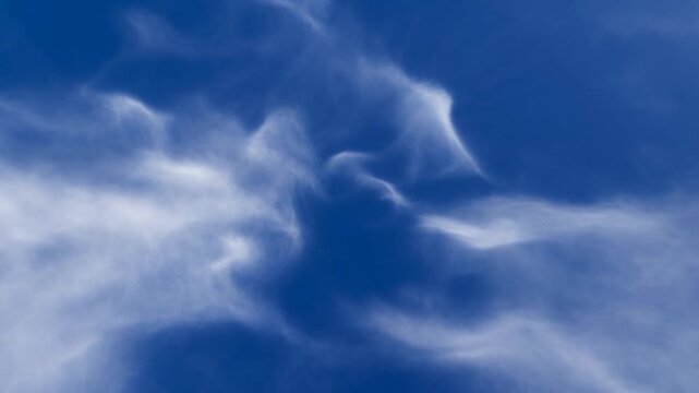 Wispy white clouds drift and change shape across an open, deep blue daytime sky