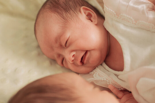 Newborn twin babies lying side by side on soft cream blanket, on sleeping peacefully and the other crying, illustrating sibling bonding, infant emotions, and early family life.