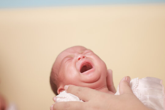 Crying newborn baby being gently supported by an adult's hands, infant emotions, parental comfort, and nurturing care. Wearing a white outfit against a warm pastel background