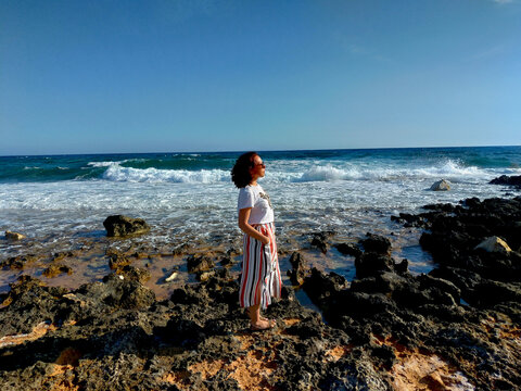 Sunlight falls across a woman and the textured rocks beside the sea, creating an informal travel portrait with open water and rolling surf