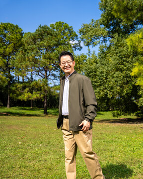 A confident, mature Chinese man, dressed in business-casual attire, stands outdoors against a backdrop of trees and a blue sky.