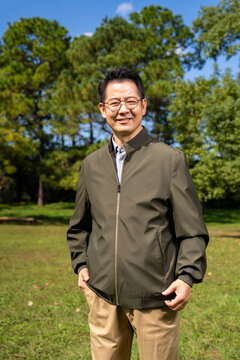A confident, mature Chinese man, dressed in business-casual attire, stands outdoors against a backdrop of trees and a blue sky.