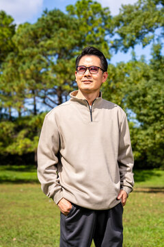A middle-aged Asian man wearing glasses stands in a park.