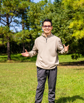 A middle-aged Asian man wearing glasses stands in a park.