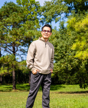 A middle-aged Asian man wearing glasses stands in a park.