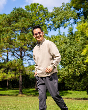 A middle-aged Asian man wearing glasses stands in a park.