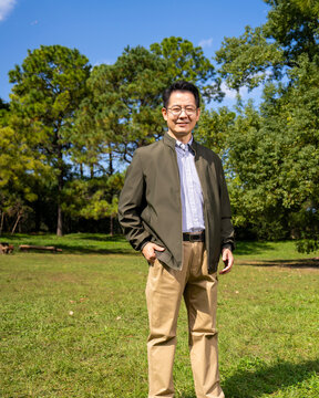 A confident, mature Chinese man, dressed in business-casual attire, stands outdoors against a backdrop of trees and a blue sky.
