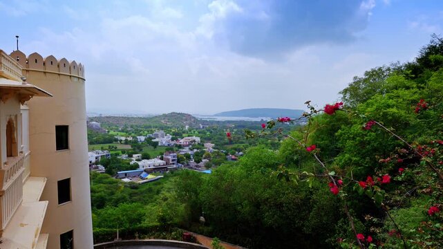 A cream building and dense flowering greenery frame a broad view across Udaipur's outer neighborhoods.
