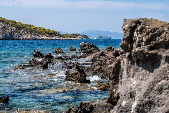 Clear turquoise waters lap against the rugged rocky shore near Perdika village on Aegina Island, offering a scenic view of Moni Island and distant sailboats in the Saronic Gulf