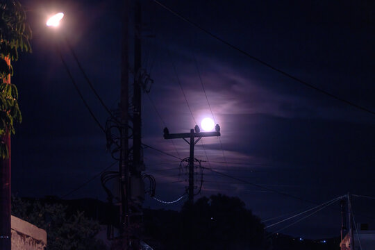 A bright full moon shines through wispy night clouds directly behind an electricity pole with power lines, illuminating the dark street and rooftops of a coastal village on Aegina Island, Greece.