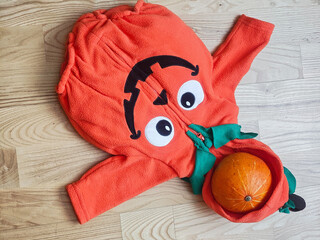 Funny Orange Halloween Costume with Pumpkin on Wooden Floor, Bright orange fleece Halloween costume featuring a funny cartoon face with large eyes and a black arrow design, green fabric details, © Алеся Макаревич