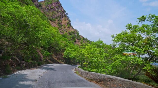 A vehicle follows a bend beside a rocky slope and low stone parapet on a wooded hill road in Udaipur.