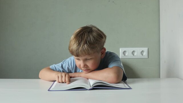 A young schoolboy sits at a desk reading an open book at home, appearing distracted and disengaged from his studies. Concept of childhood education, reading difficulties, homework and concentration.