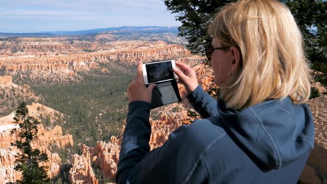 Blonde female traveler standing on cliff edge taking photos with smartphone at bryce canyon national park, capturing red rock hoodoo formations and scenic wilderness on sunny day