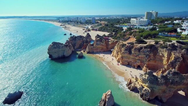 Drone camera flying over praia dos tres irmaos in algarve portugal, revealing turquoise ocean waters, golden sandstone cliffs, sandy beaches, and resorts under a clear blue sky