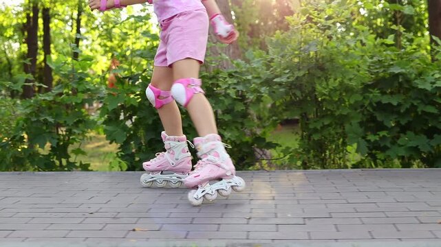 Close up of child legs in pink inline skates and knee pads riding along a alley in a summer park in slow motion. Active kids outdoor leisure concept. Rollerblading.
