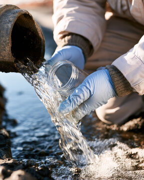 Industrial water discharge sampling with gloved environmental worker filling sterile glass jar beside drainage pipe in sunlit canal
